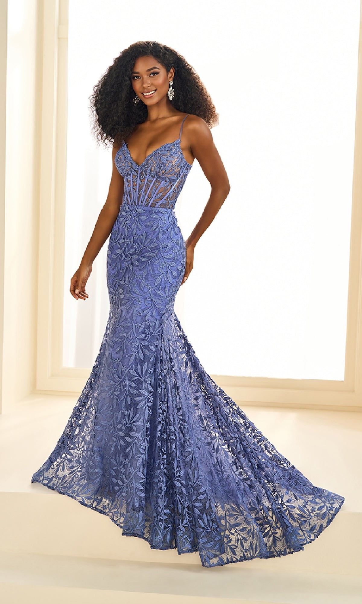 A woman stands in a well-lit room wearing the Ellie Wilde EW36011, a floor-length fitted lavender lace wedding dress with spaghetti straps and a flared hem.